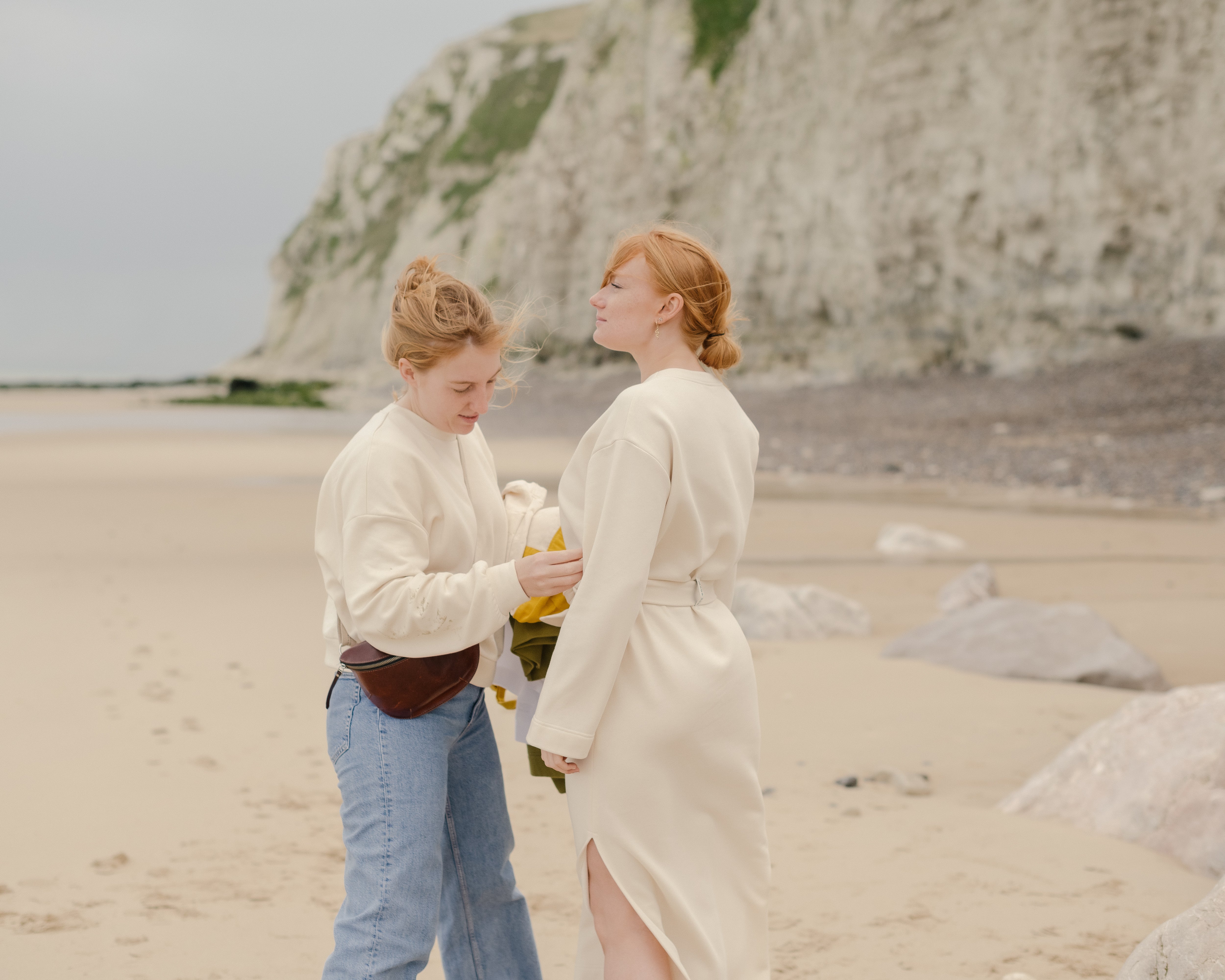 Two girls by the seaside in soft-toned clothes, one adjusting the belt of the other's dress.